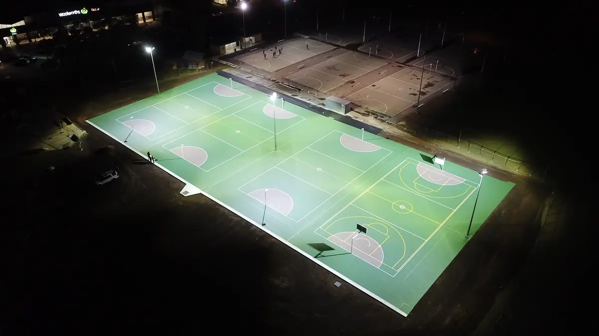 Floodlit multi-sport netball and basketball courts at night, with the old cracked courts visible alongside