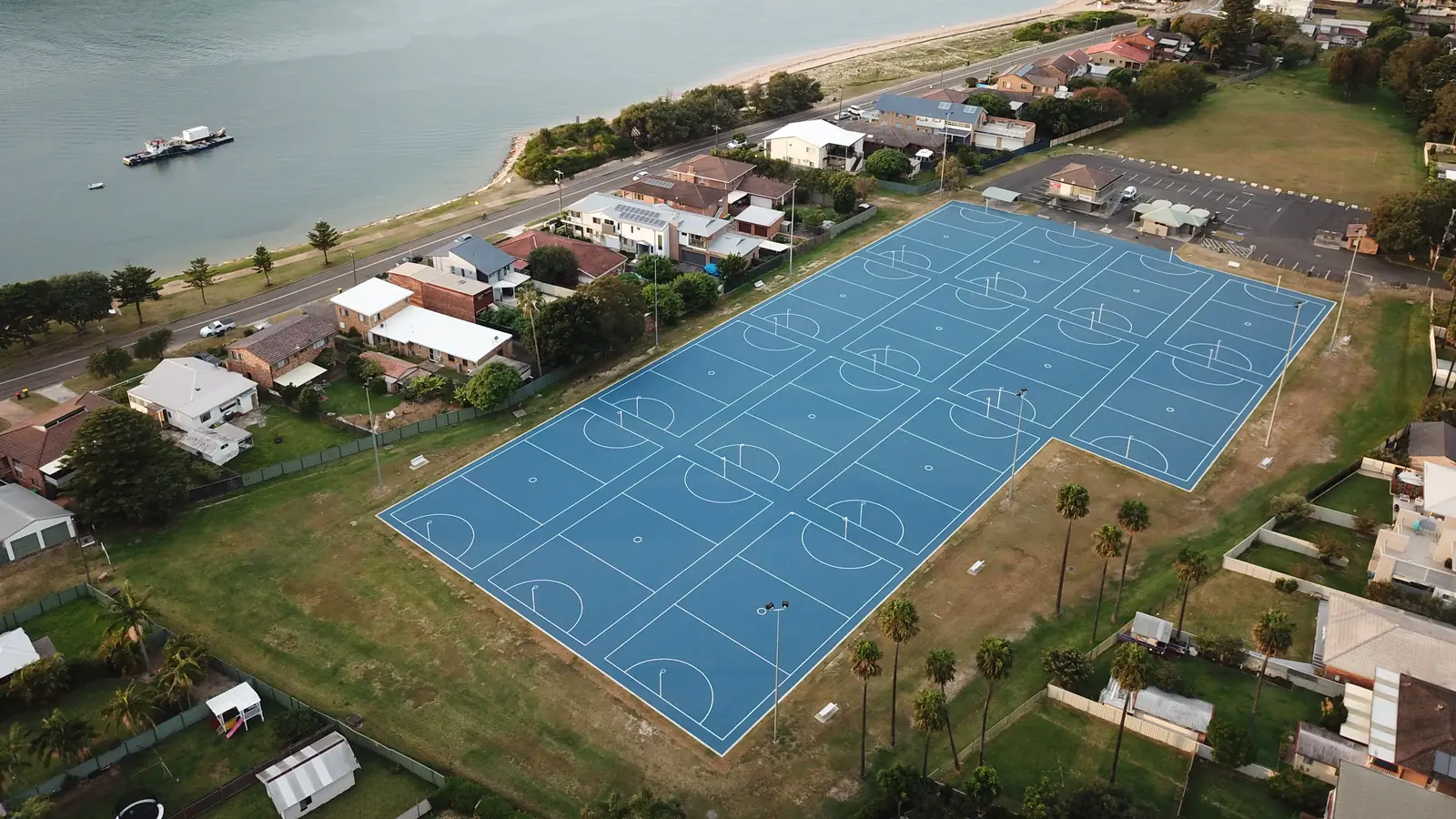 Aerial view of a completed multi-court blue netball complex beside a waterway, with a beach, headland and surrounding houses behind it